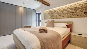 Quarto moderno com cama grande, cabeceira de madeira, parede de pedra e mesa de cabeceira minimalista.