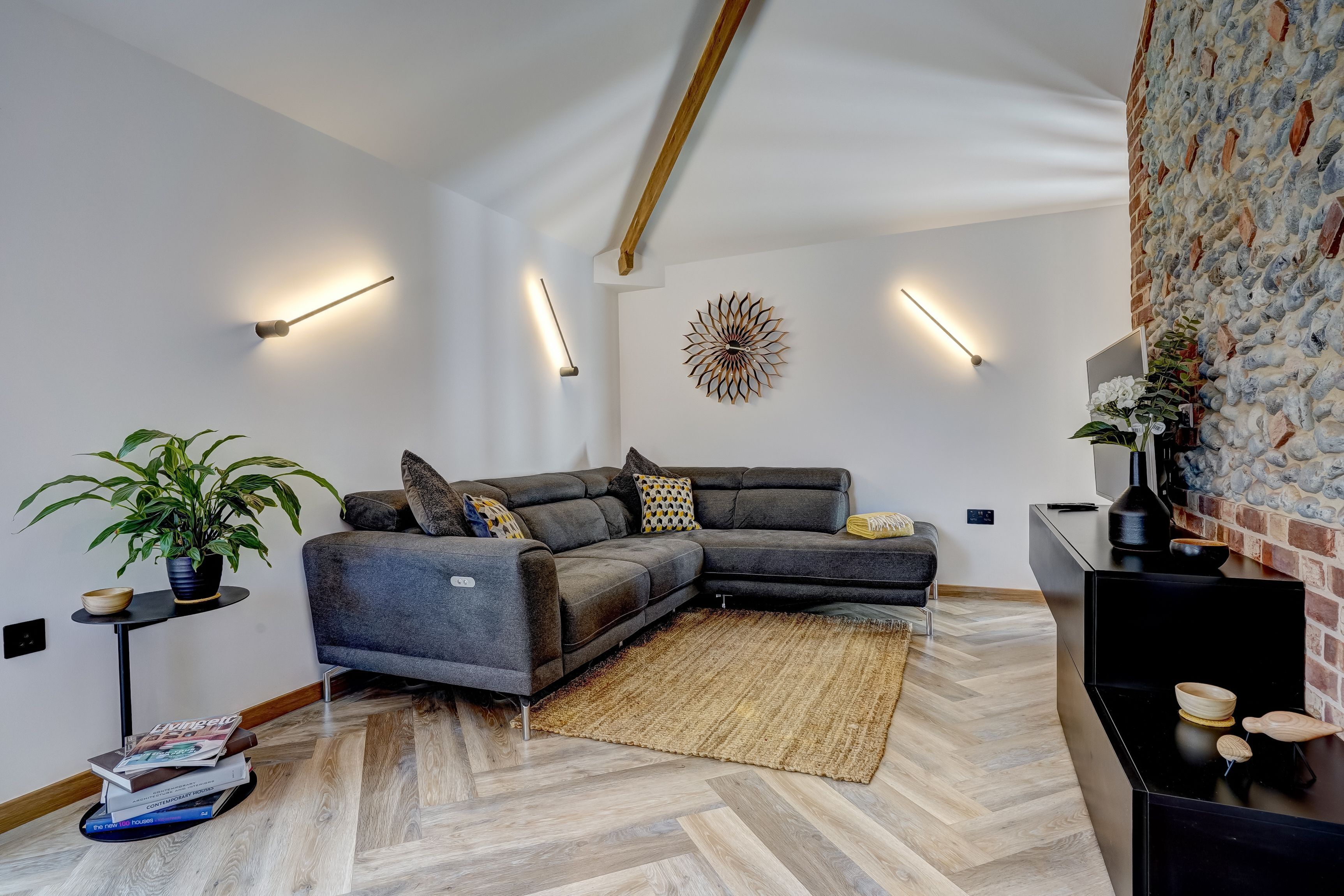 A modern living room with a sectional sofa, a herringbone-patterned floor, a decorative wall piece, and a small side table with a plant.