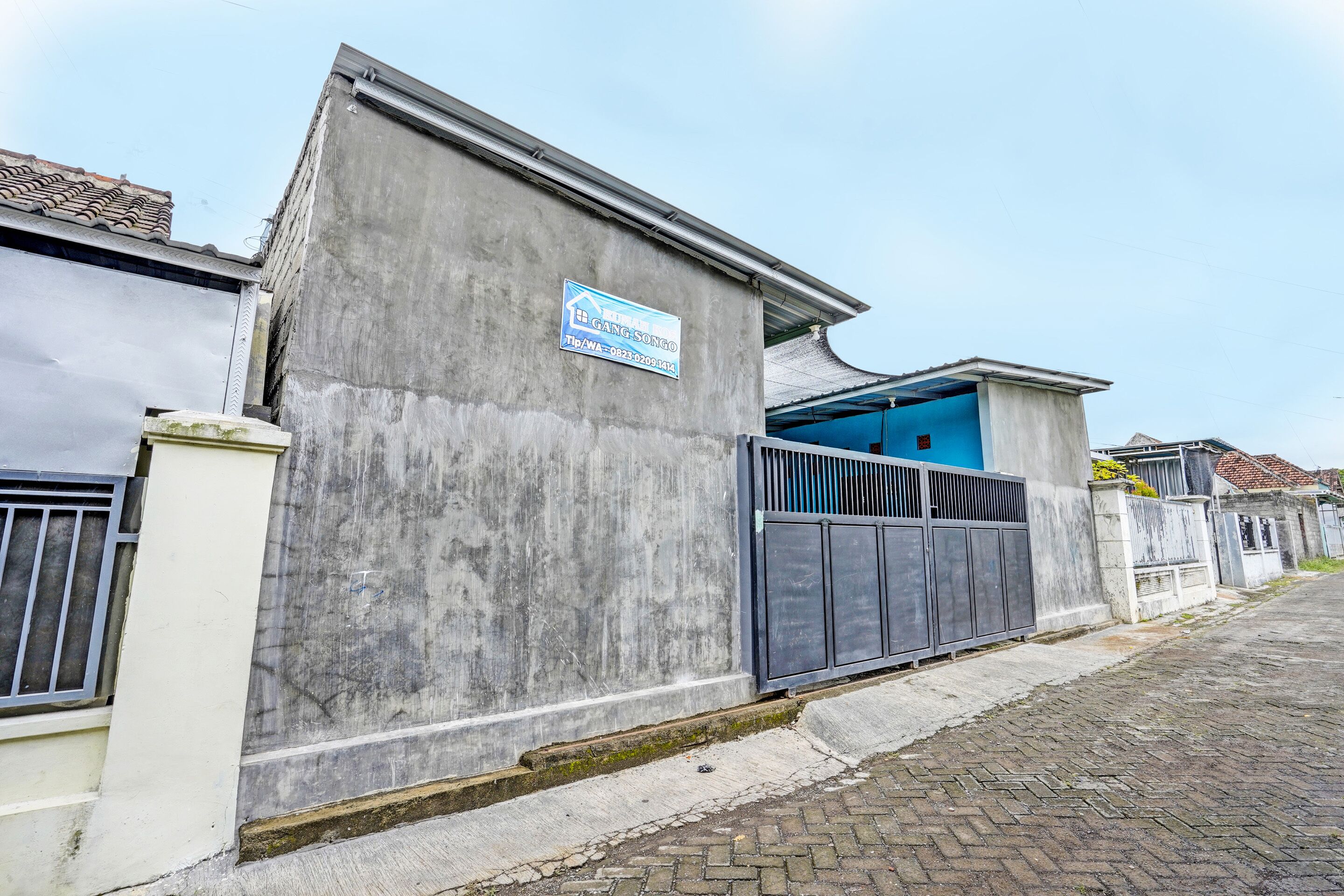 Un edificio de hormigón con una puerta de garaje azul, entrada protegida por una cancel y un letrero que indica un negocio llamado 'GANG SONGO'.