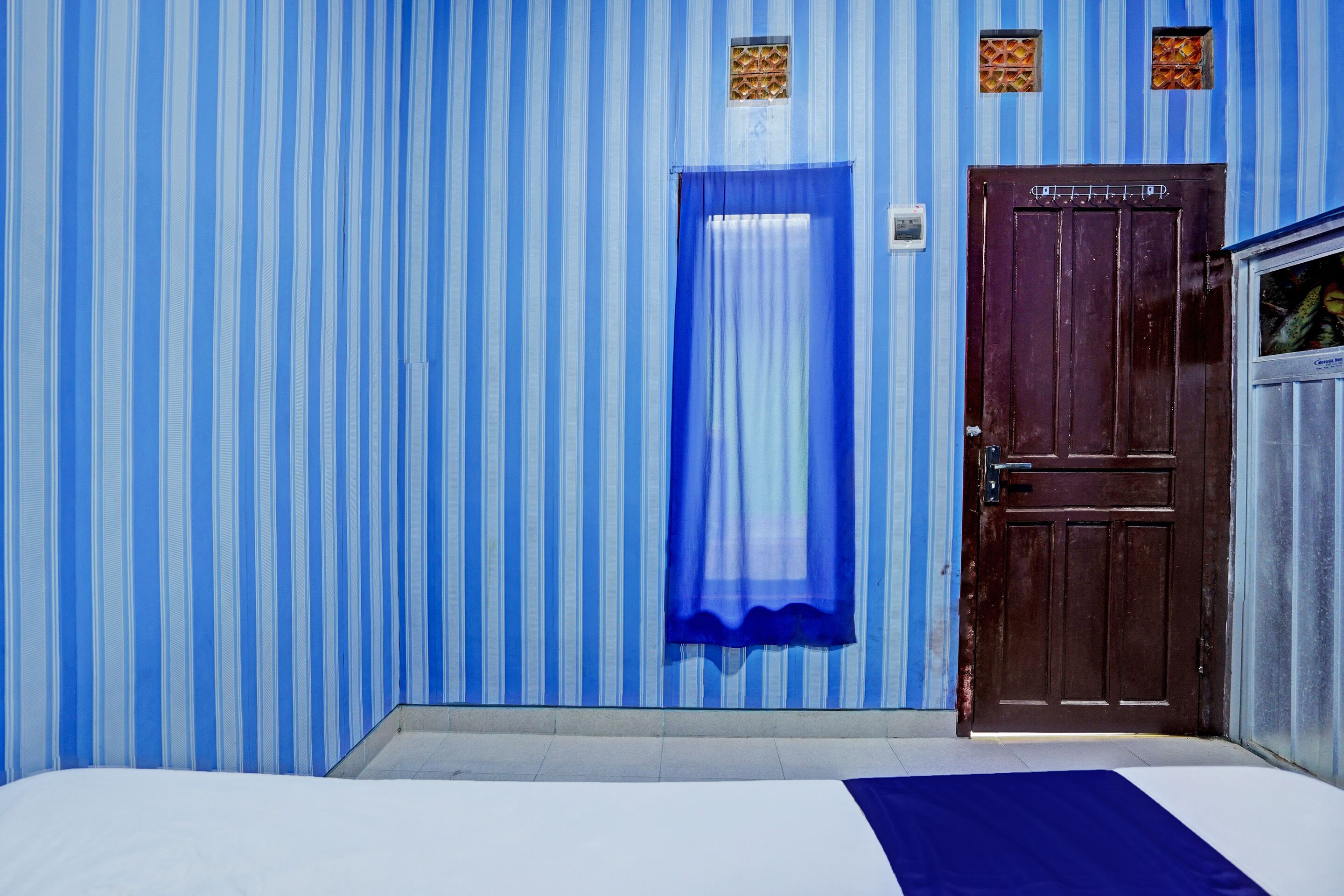 A room with a bed, a blue-striped wall, a wooden door, and a window with a blue curtain.