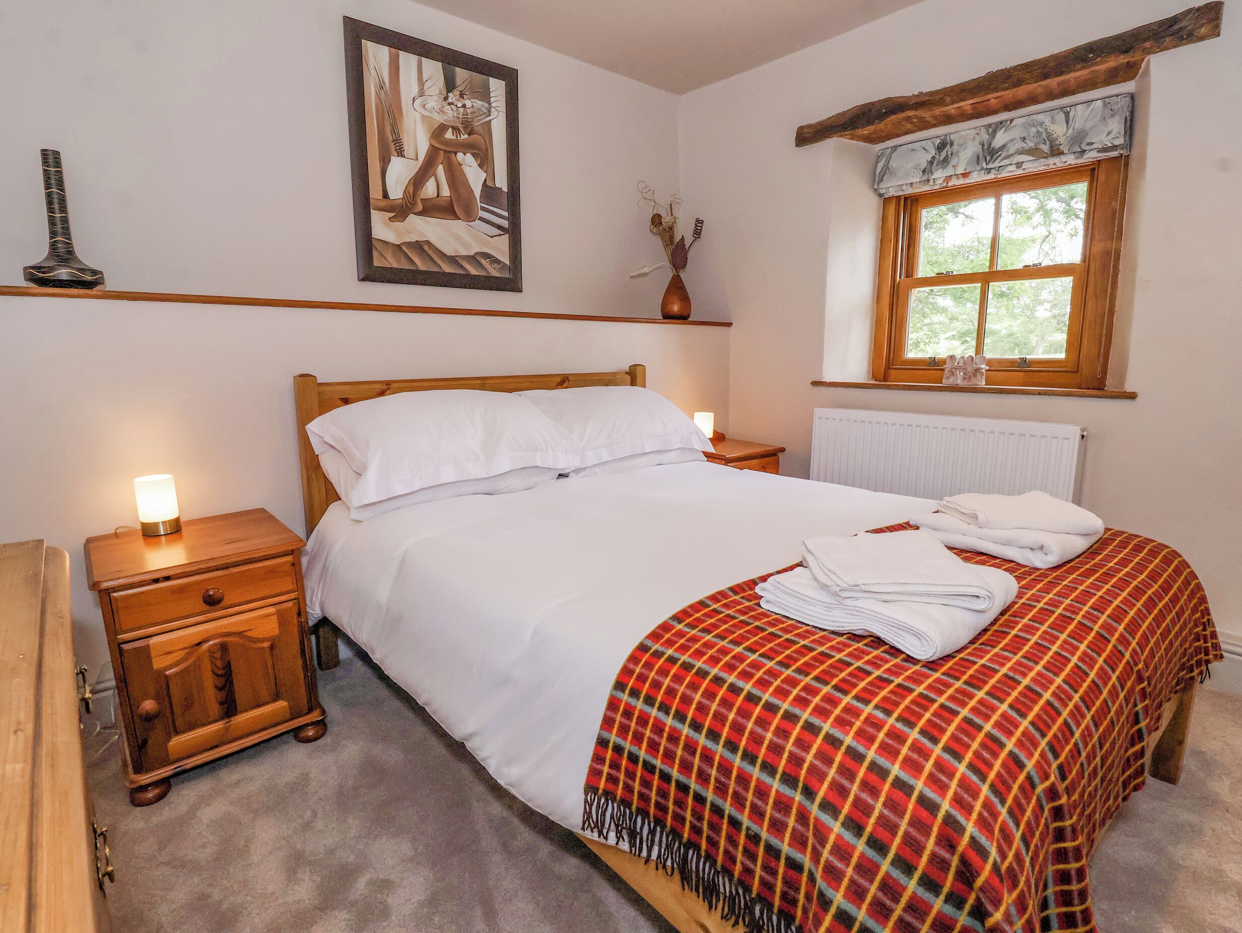 A neatly made bed with white linens and a plaid blanket, a bedside table with a lamp, a framed artwork on the wall, and a window with a view of trees.