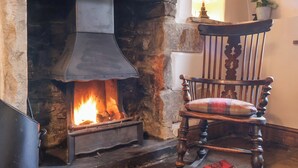 Une cheminée en pierre avec un feu de bois, un fauteuil en bois avec un coussin à carreaux et une lampe posée sur une étagère.
