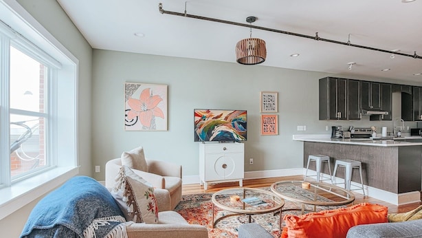 Una sala moderna con sofá, sillón y mesa de centro, una cocina con alacenas oscuras y obras de arte en las paredes.