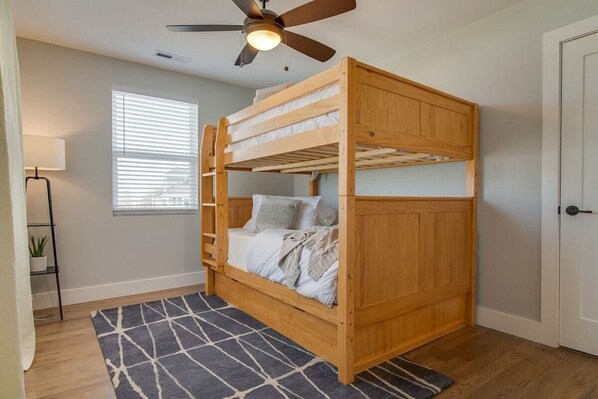 A bunk bed with a ceiling fan, a window with blinds, a bedside table with a lamp, and a patterned rug.