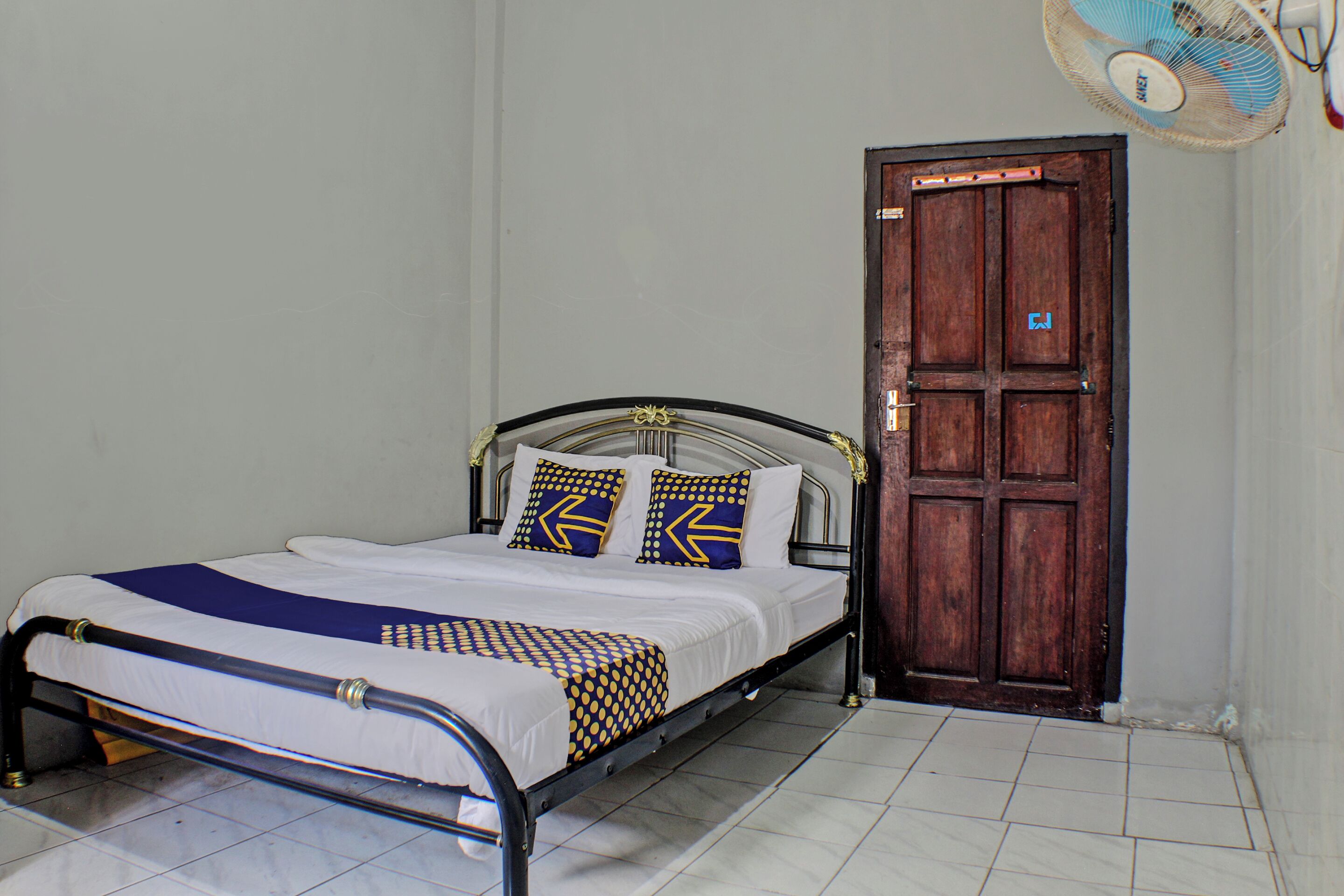 A single bed with a metal frame, white bedding, and patterned pillows. A wooden door is visible to the right, and a ceiling fan is mounted above the bed.