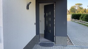 Ein moderner Gebäudeeingang mit einer dunklen Tür, ein kleiner Außenbereich mit Fußmatte und ein gepflasterter Weg, der zu einem Zaun führt.