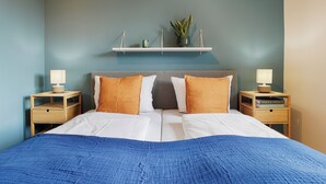 En pænt redt seng med hvidt og blåt sengetøj, to sengeborde med lamper, og en hylde over sengen med en plante og en lille genstand.