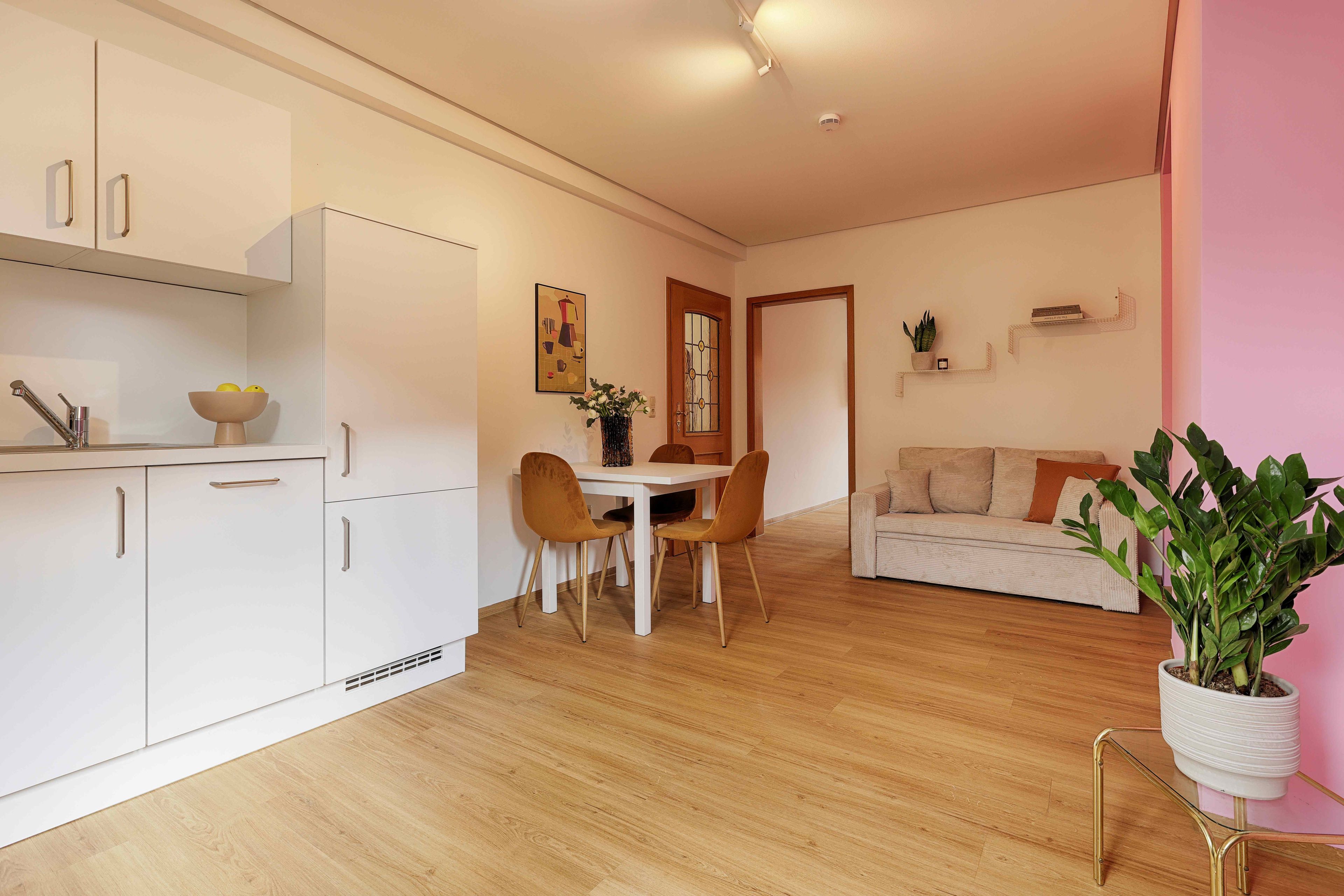 A modern kitchen with white cabinets, a dining table with two chairs, and a living area with a sofa and a potted plant.
