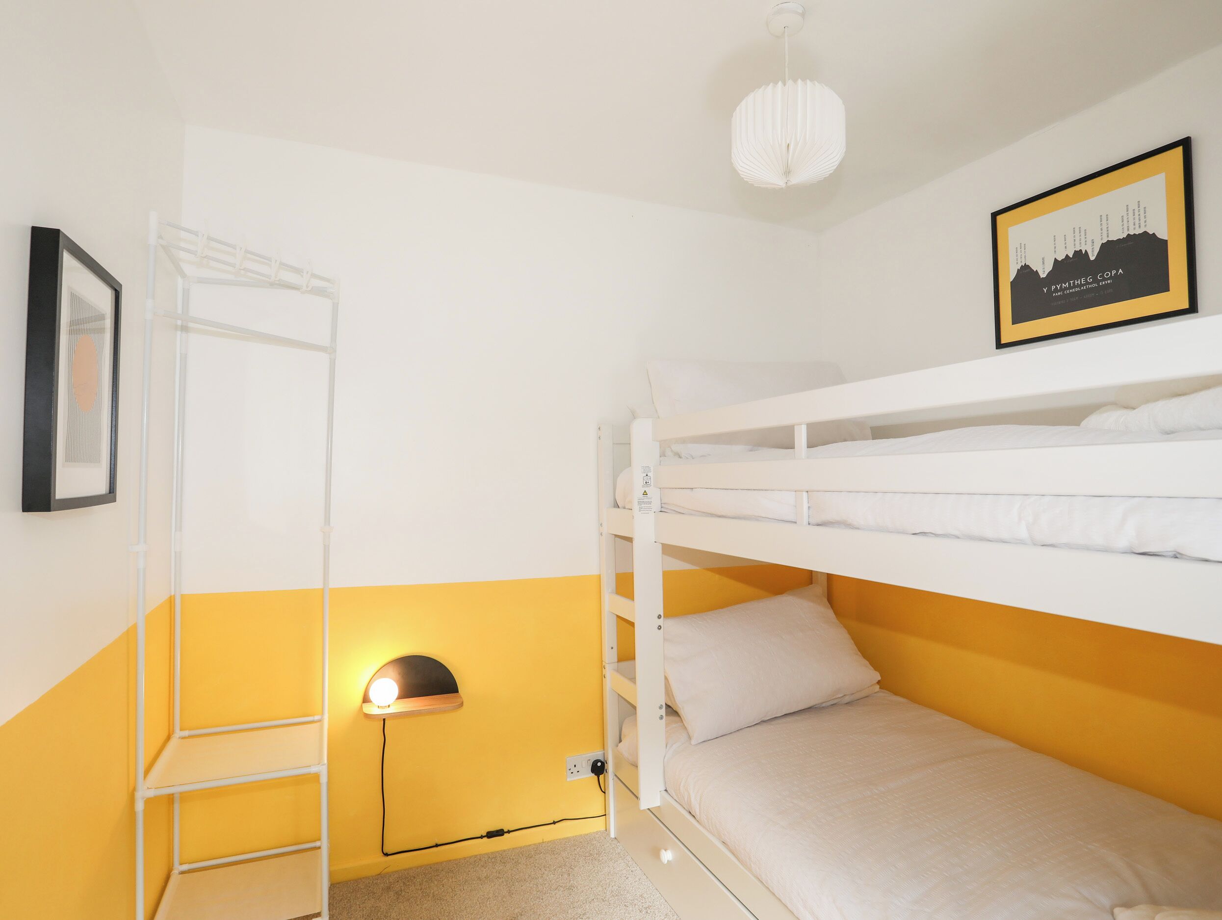 A bunk bed room with a white and yellow color scheme, a framed picture on the wall, and a hanging light fixture.