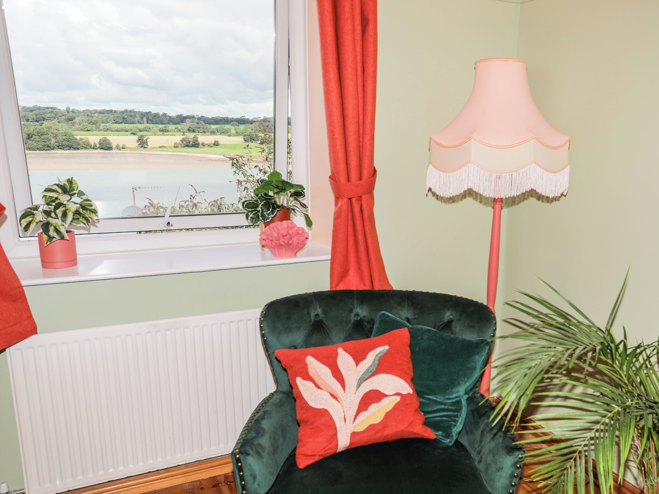 Una habitación con vistas a un lago, una cortina roja, una lámpara rosa y una silla verde con un cojín rojo.