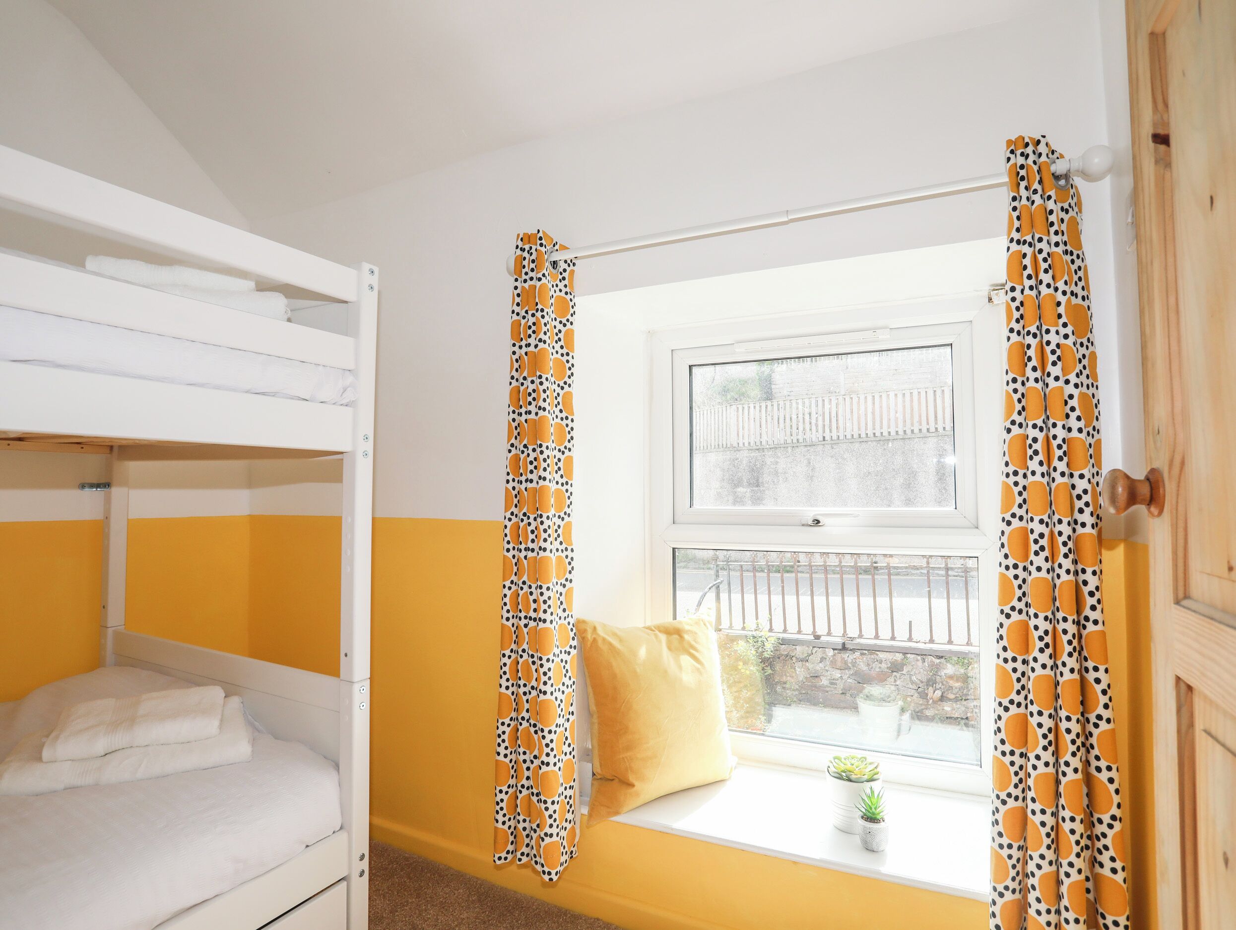 A room with a bunk bed, a window with a view of a fence and a stone wall, and a curtain with a polka dot pattern.