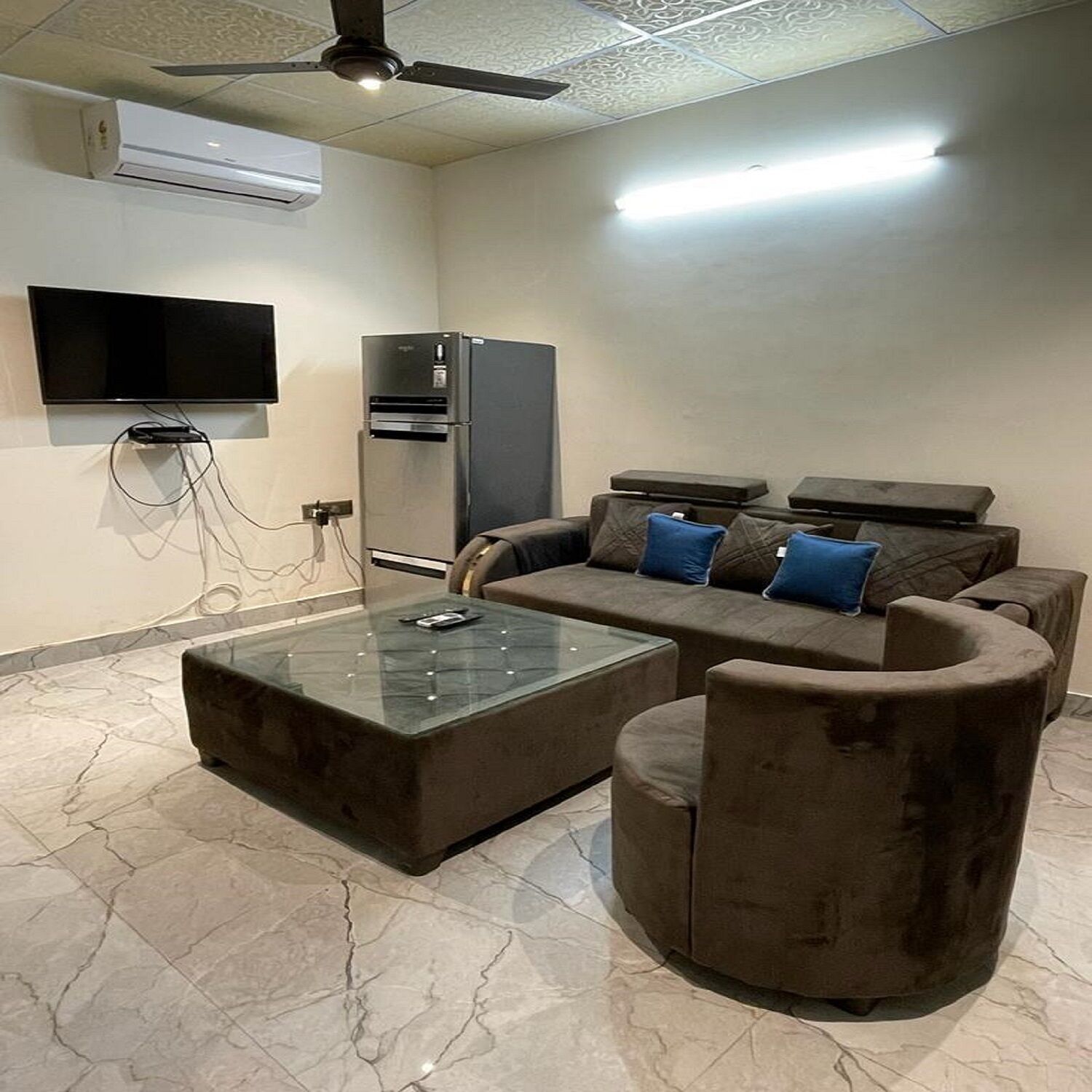 Un salon moderne avec un canapé d’angle, une table basse en verre, un téléviseur à écran plat fixé au mur et un système de climatisation intégré.