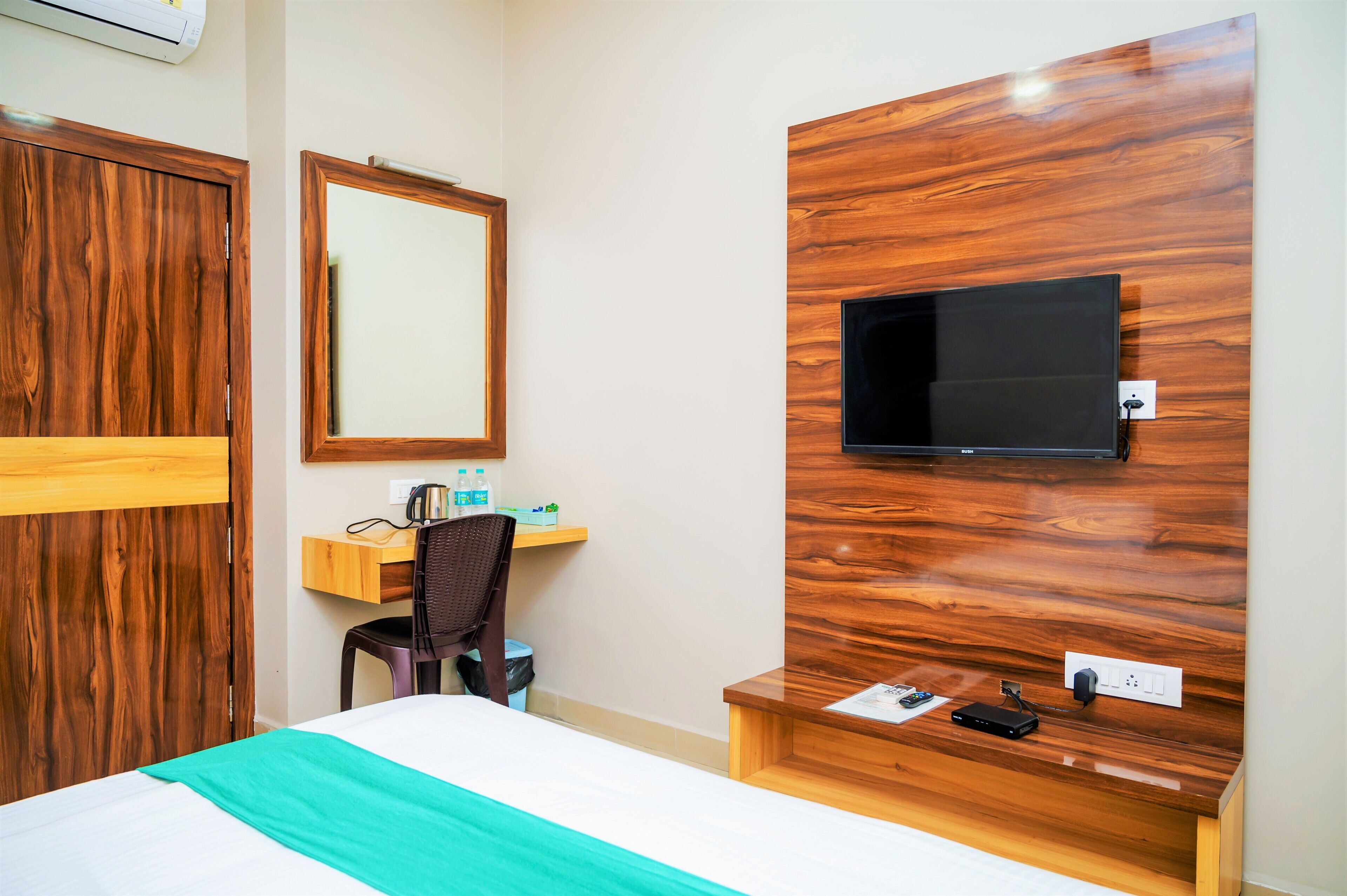 A hotel room with a bed, a wooden desk with a chair, a mirror, a television mounted on a wooden wall, and a small shelf with toiletries.