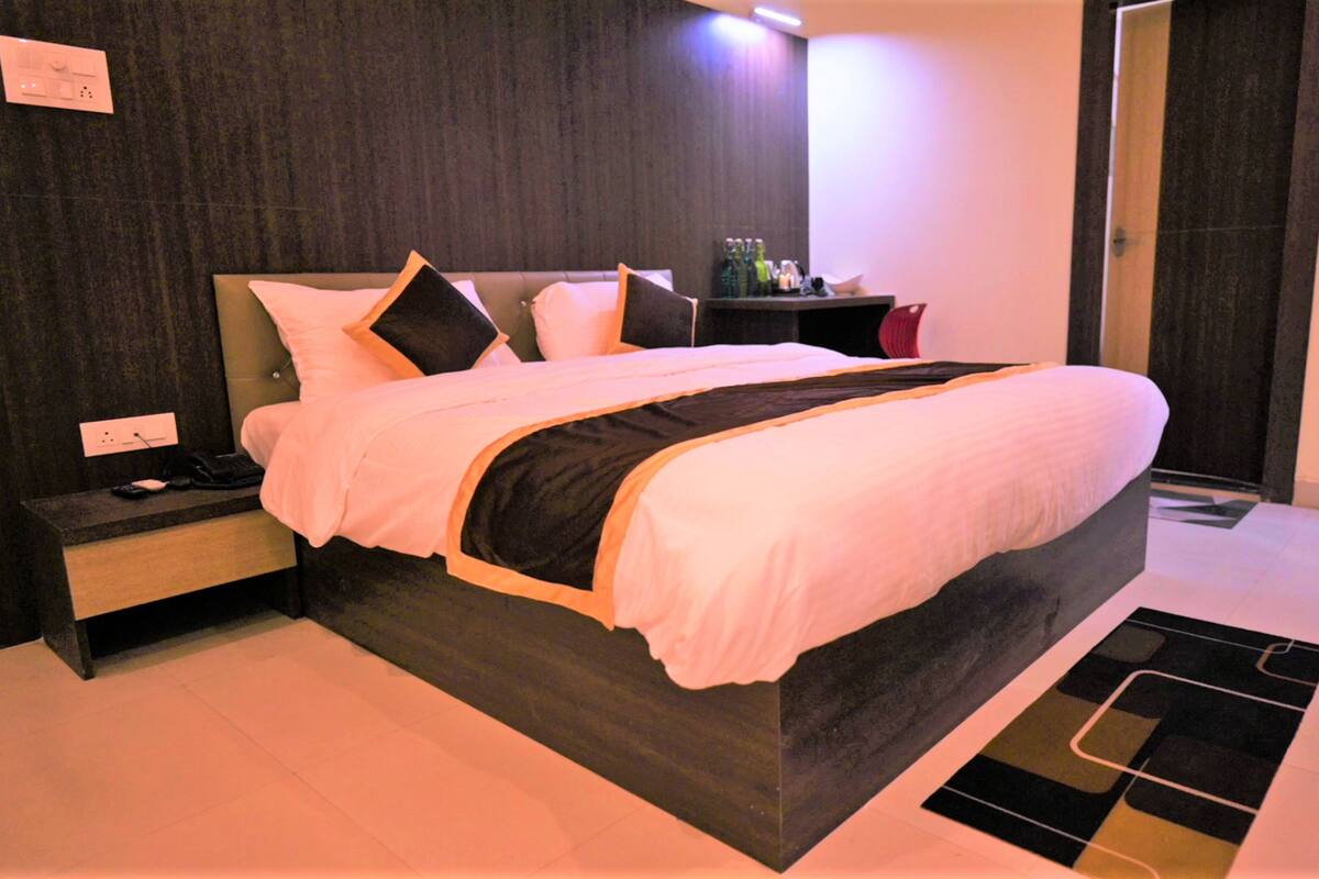 A modern hotel room with a large bed, a bedside table, a nightstand, and a small shelf with bottles.