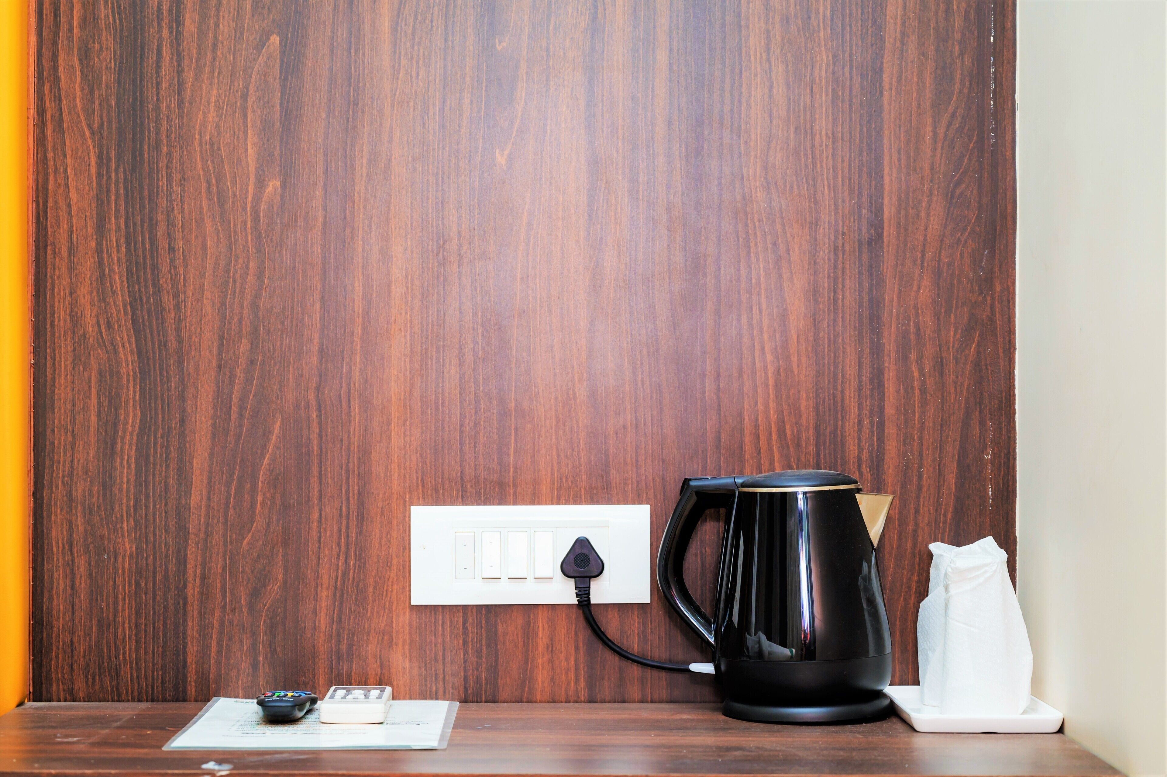 A wooden wall with a power outlet, a black coffee maker, a remote control, and a white bag on a wooden table.