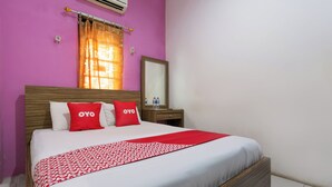 Quarto de hotel com uma cama grande, duas almofadas com a marca 'OYO', uma cabeceira de madeira, uma mesinha de cabeceira com espelho e uma janela com cortinas laranja.