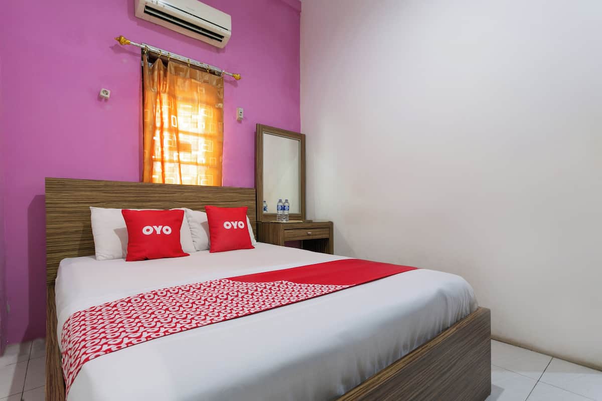 A hotel room with a large bed, two pillows with 'OYO' branding, a wooden headboard, a nightstand with a mirror, and a window with orange curtains.