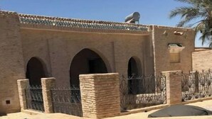 Une piscine à l’eau d’un bleu cristallin, entourée d’un bâtiment en briques doté d’arcades et, au fond, d’un palmier.