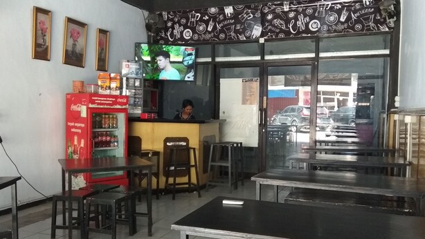 Interior de uma cafeteria com mesas, cadeiras, um refrigerador de Coca-Cola e uma televisão mostrando uma pessoa.