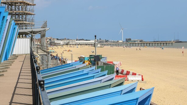Une plage avec une jetée, une éolienne et une promenade en bois bordée de bâtiments aux toits bleus.