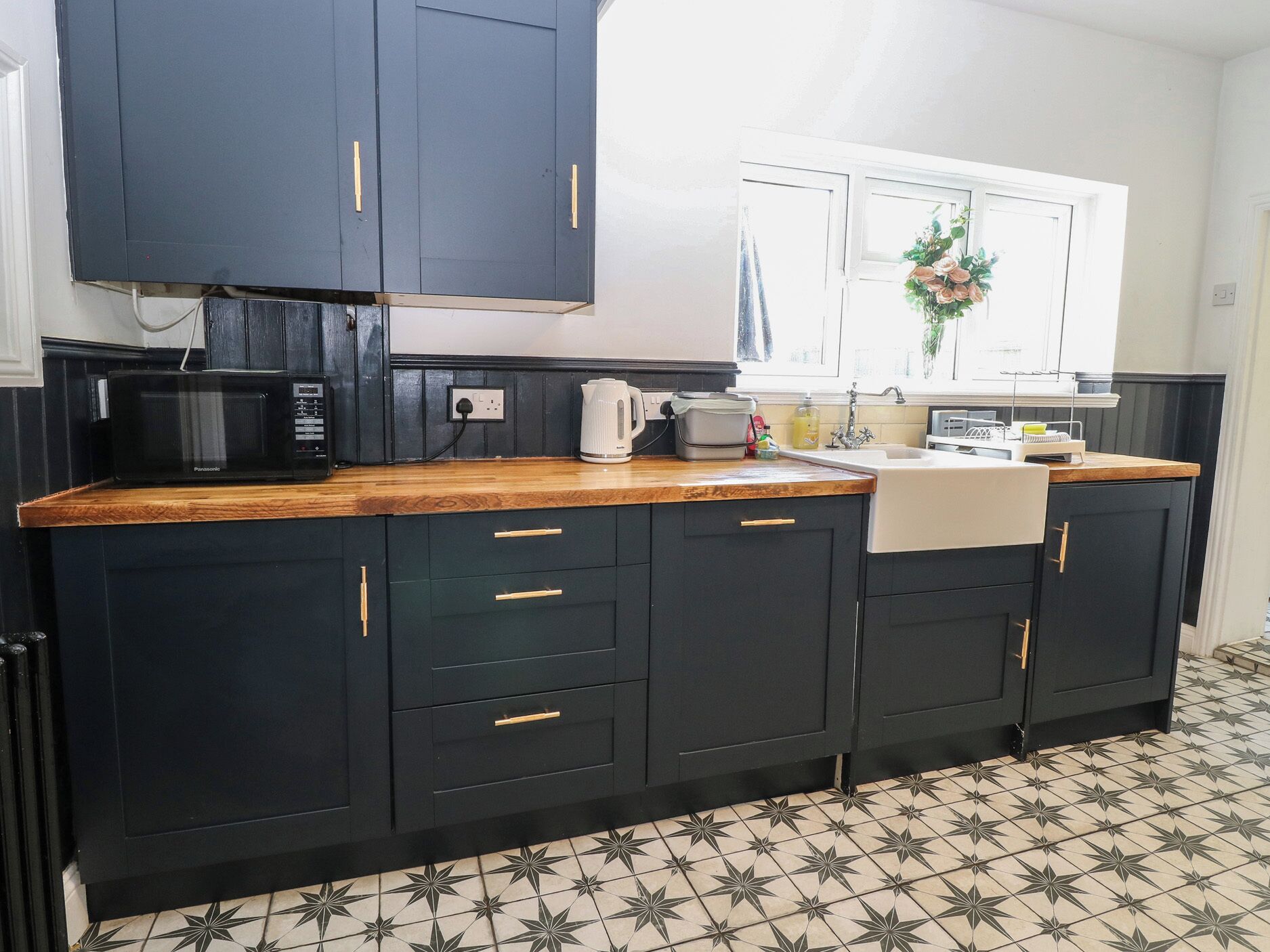A kitchen with dark cabinets, a microwave, a kettle, and a window with a flower arrangement.