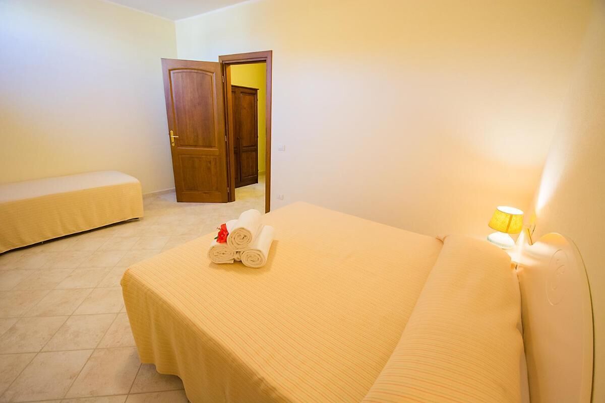 A hotel room with a bed, a bench, a door, and a lamp.