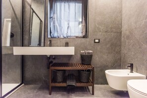 Een moderne badkamer met een marmeren wand, een wit wastafelmeubel, een bidet, een houten plank met manden, en een spiegel boven het wastafelmeubel.