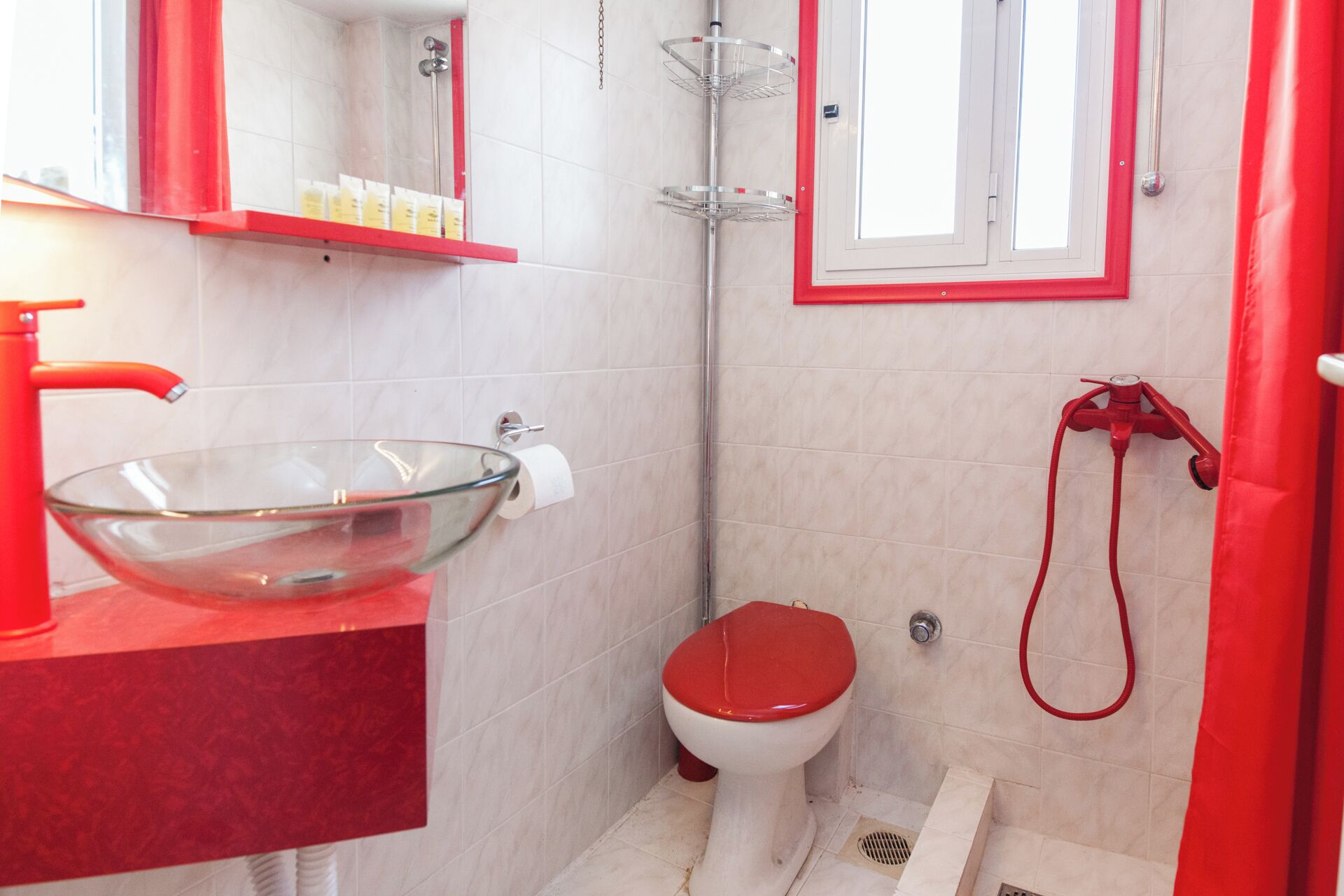 A bathroom with a glass bowl sink, red toilet, and a shower with a red hose.