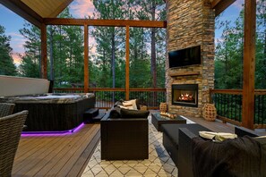 A covered outdoor area with a stone fireplace, seating, and a hot tub. - New! Peak-A-Boo Pines - Game Loft, Hot Tub, Fireplaces, Slide, Free Firewood! (Broken Bow)