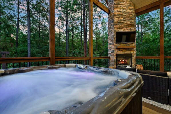 A hot tub with a stone fireplace, surrounded by wooden deck and forest views.