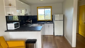 Una cucina compatta con mobili bianchi, un piano di lavoro nero, un forno a microonde, un lavandino e un frigorifero. In primo piano, una sedia gialla e un cestino per i rifiuti.