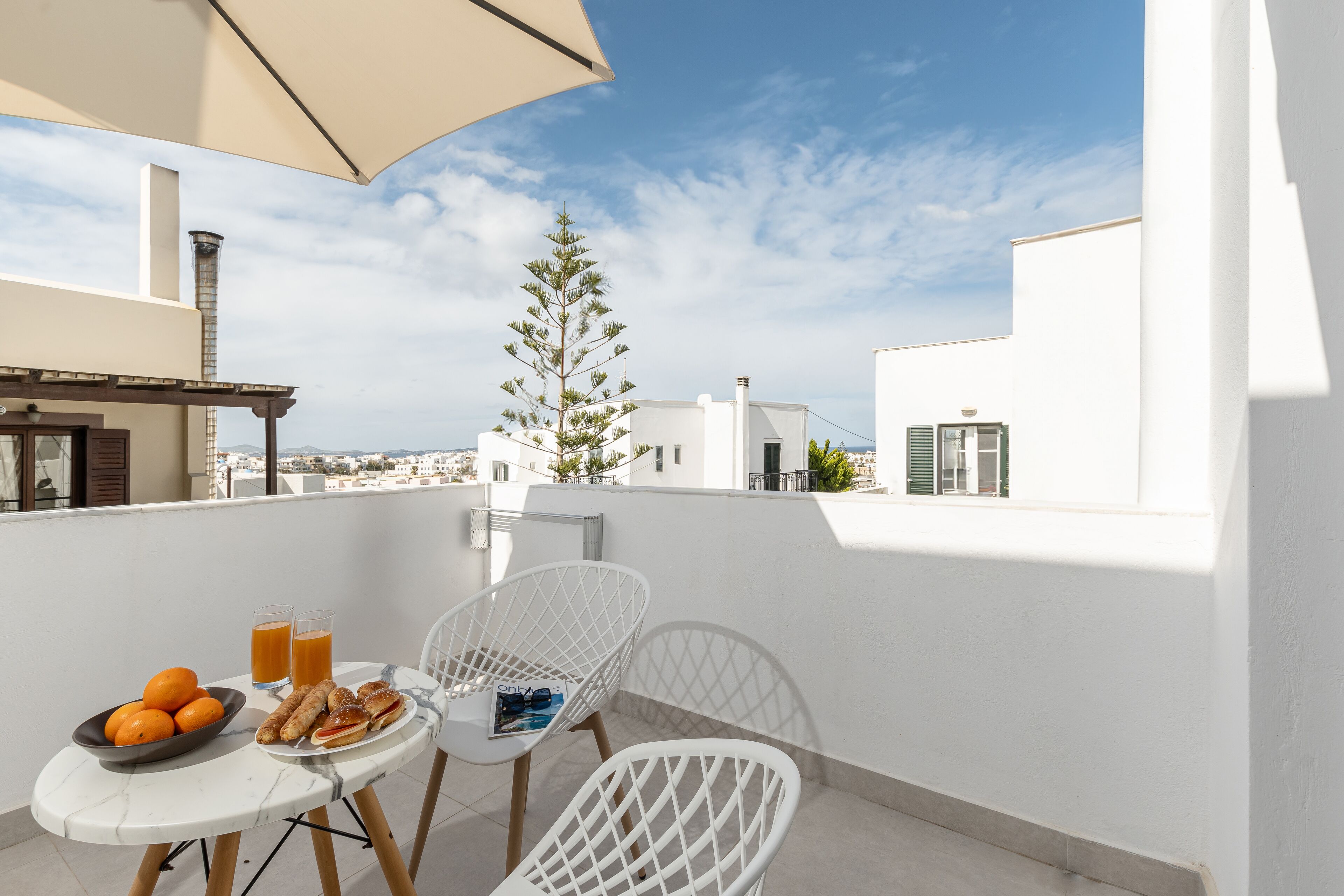 A balcony with a table set for breakfast, including oranges and pastries, and two white chairs.