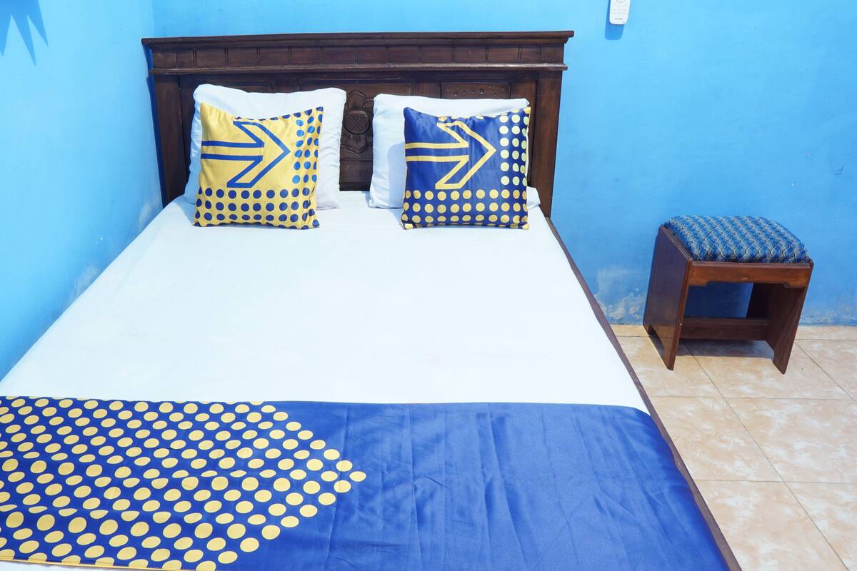 Una cama con dos almohadas, una colcha de lunares azules y amarillos, y una mesita de noche de madera con un cojín azul.