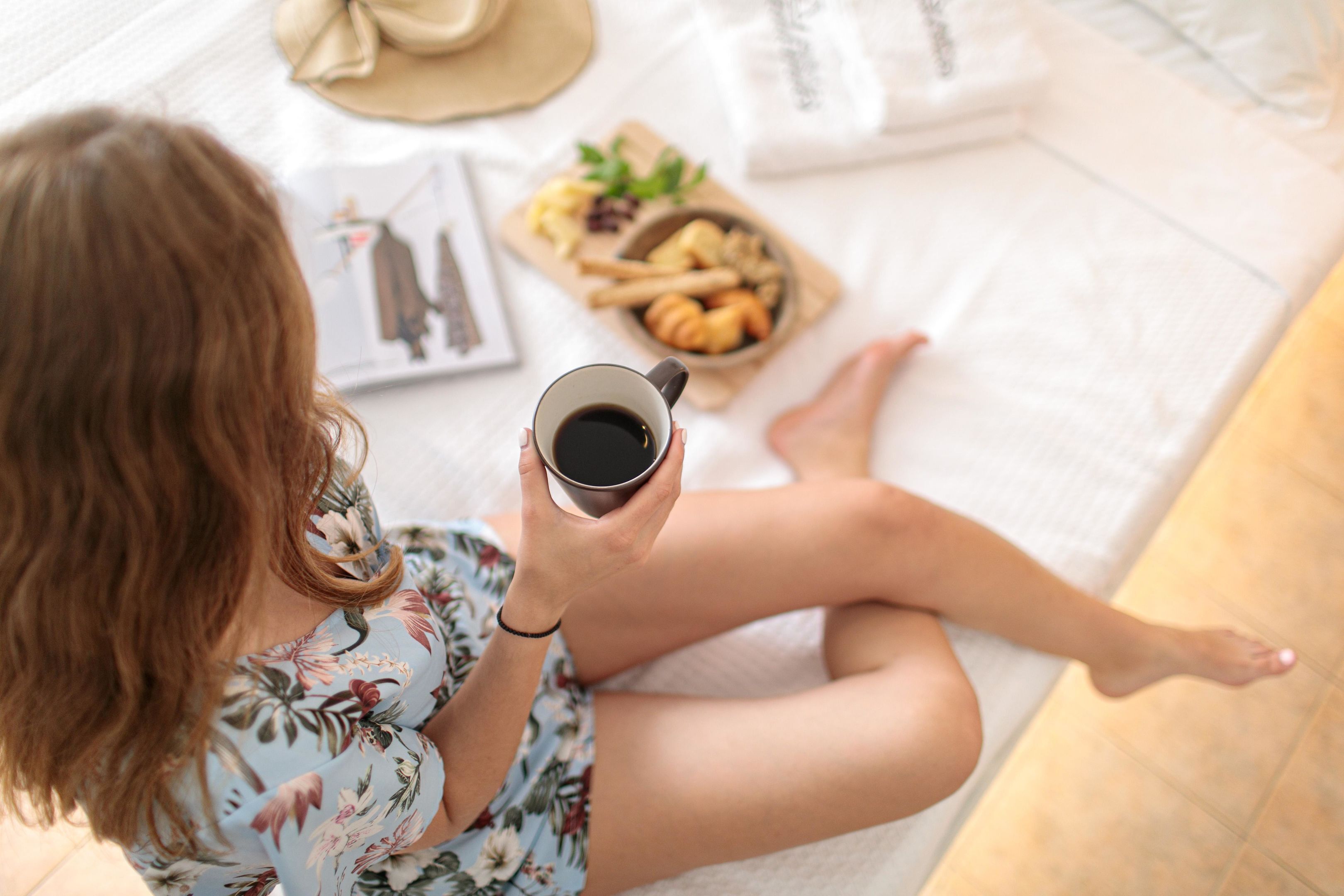 Eine Person sitzt auf einem Bett mit einem Buch, einer Kaffeetasse und einem Tablett Gebäck.