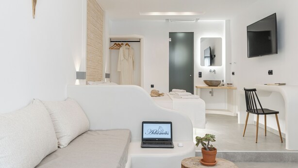 A modern hotel room with a sofa, a laptop, a potted plant, a wooden chair, a mirror, and a TV mounted on the wall. - Adama's Suites Naxos (Naxos)