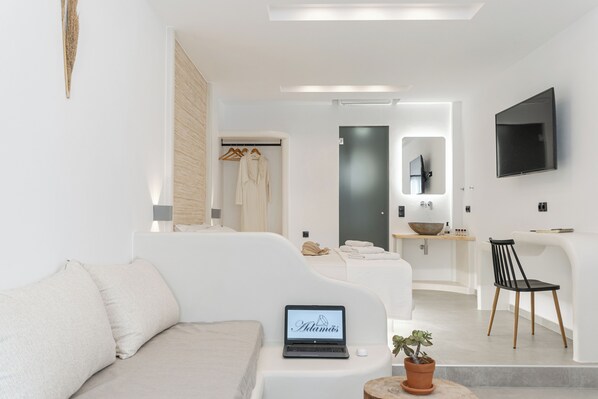 A modern hotel room with a sofa, a laptop, a potted plant, a wooden chair, a mirror, and a TV mounted on the wall.