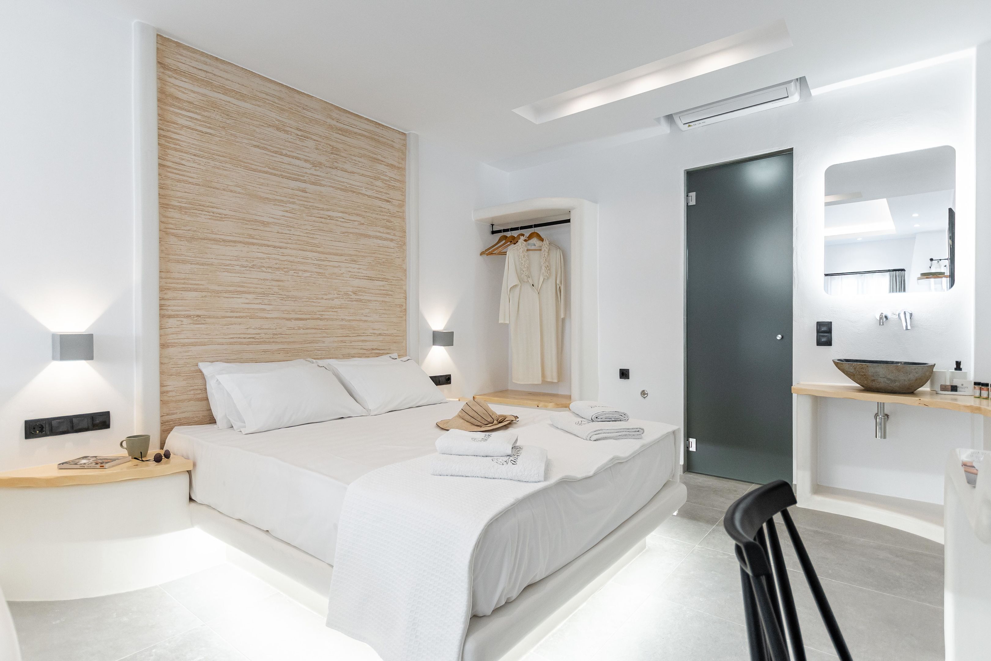 A modern hotel room with a large bed, bedside tables, a wooden accent wall, a bathroom with a sink and mirror, and a door leading to another room.