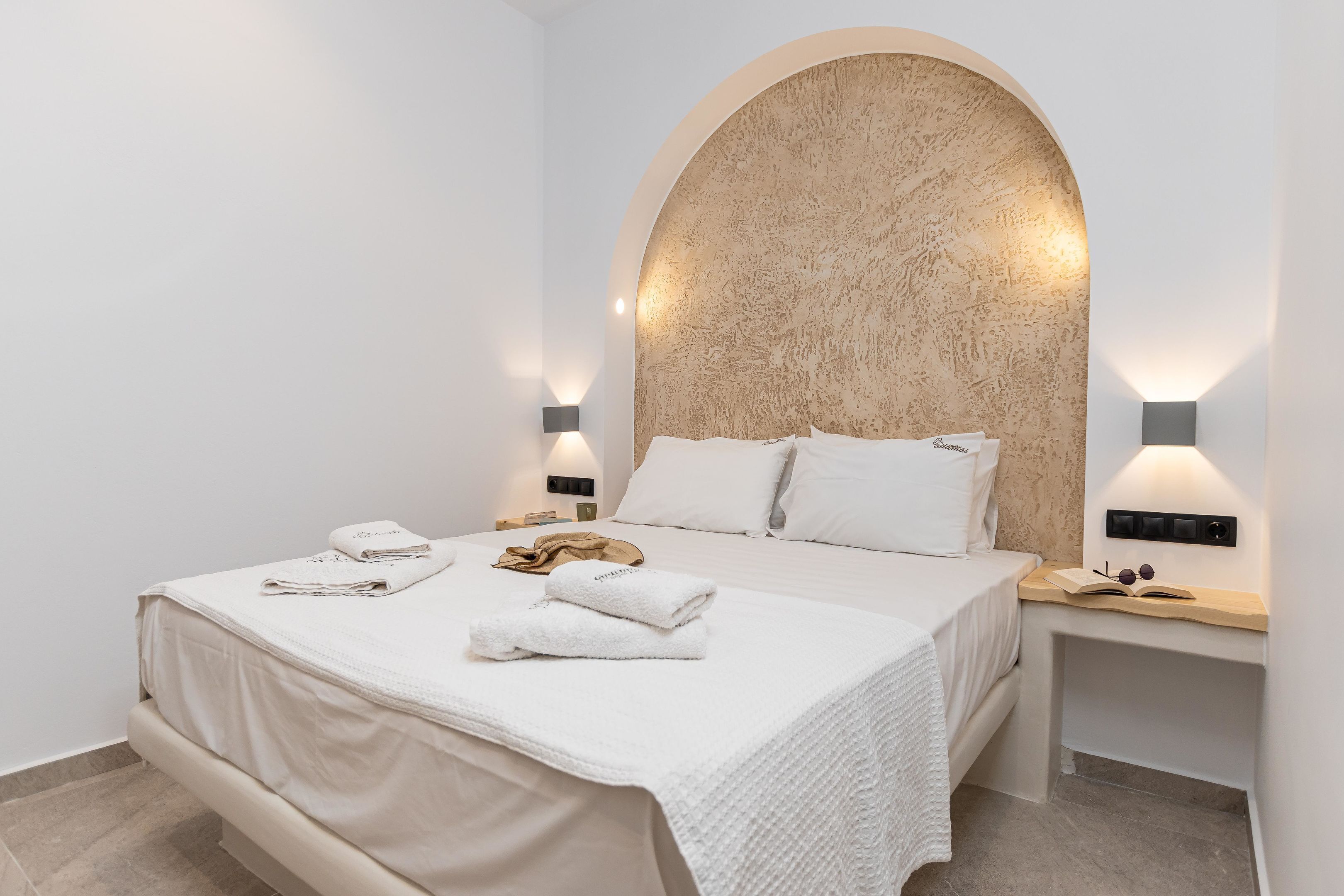 Un dormitorio moderno con un gran cabecero arqueado, una cama bien tendida con sábanas blancas y dos mesitas de noche con lámparas de pared.