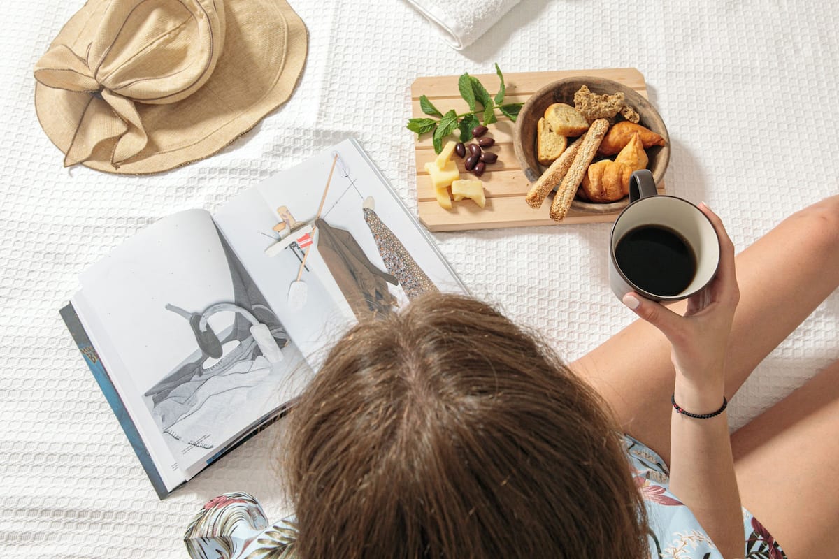 A person lying on a bed with a book, a hat, a tray of food, and a cup of coffee.