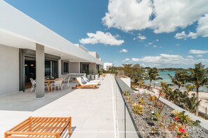A modern outdoor terrace with a dining area, lounge chairs, and a view of the beach and ocean.
