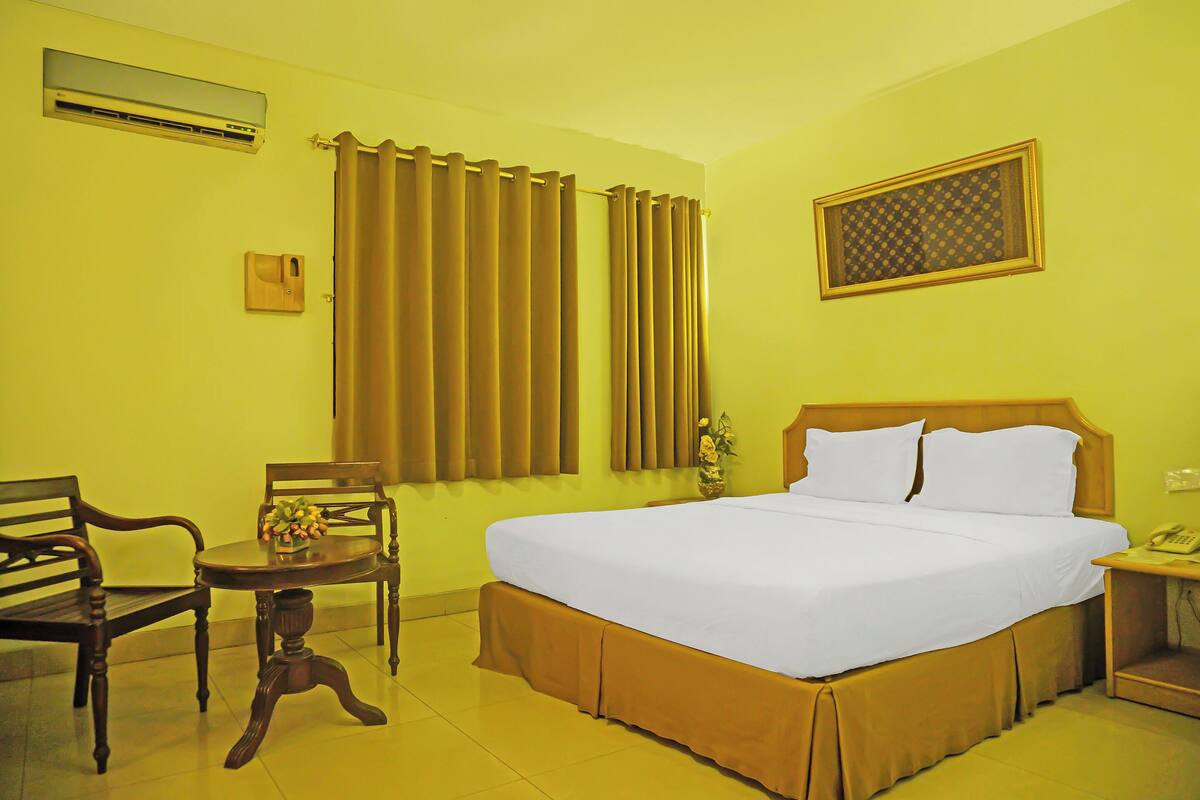 Quarto de hotel com uma cama grande, uma cadeira de madeira, uma mesa pequena com um vaso e uma janela com cortinas.