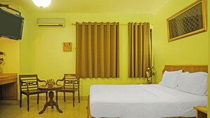 Une chambre d’hôtel avec un lit, deux chaises, une petite table ornée de fleurs, un appareil de climatisation, des rideaux, un téléviseur et un tableau encadré accroché au mur.