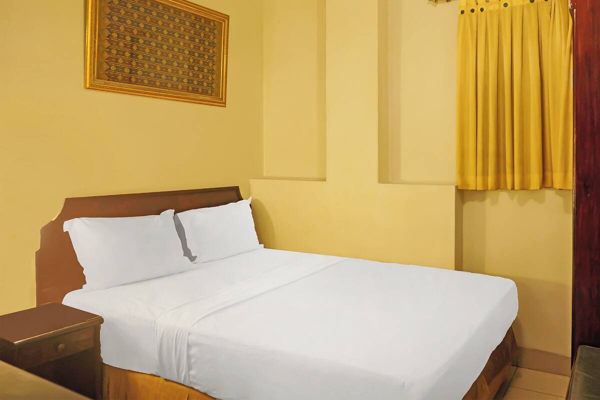 Uma cama bem arrumada com lençóis brancos, cabeceira de madeira e mesa de cabeceira com gaveta.