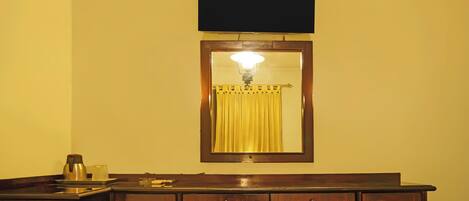 A hotel room with a wooden desk, a flat-screen TV mounted above it, and a mirror with a light fixture.