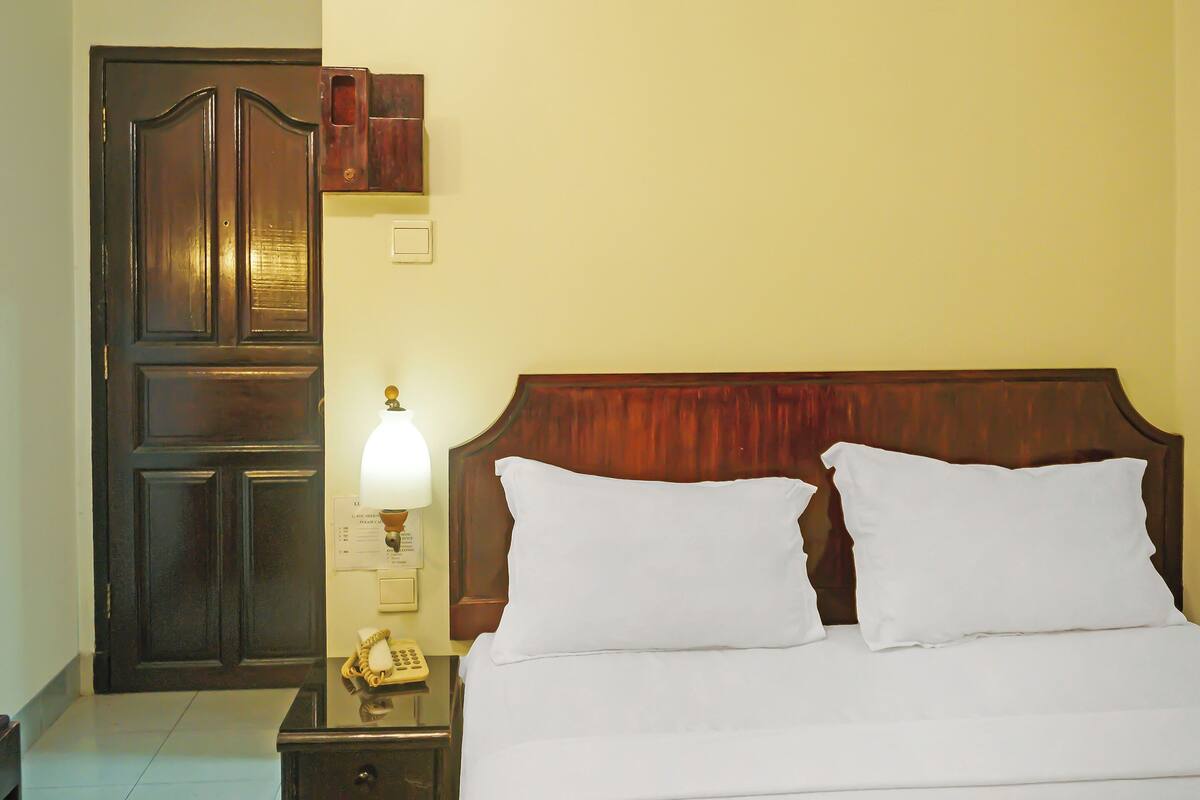 Quarto de hotel com porta de madeira, mesa de cabeceira com abajur e cama com roupa de cama branca.