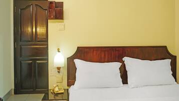 Habitación de hotel con puerta de madera, mesita de noche con lámpara y cama con ropa blanca.