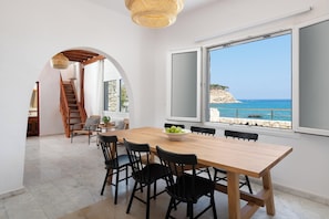 A dining area with a wooden table and chairs, a view of the sea, and a staircase leading to another room.