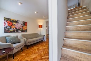 A modern living room with a herringbone wooden floor, two grey sofas, a painting of flowers, and a wooden staircase.