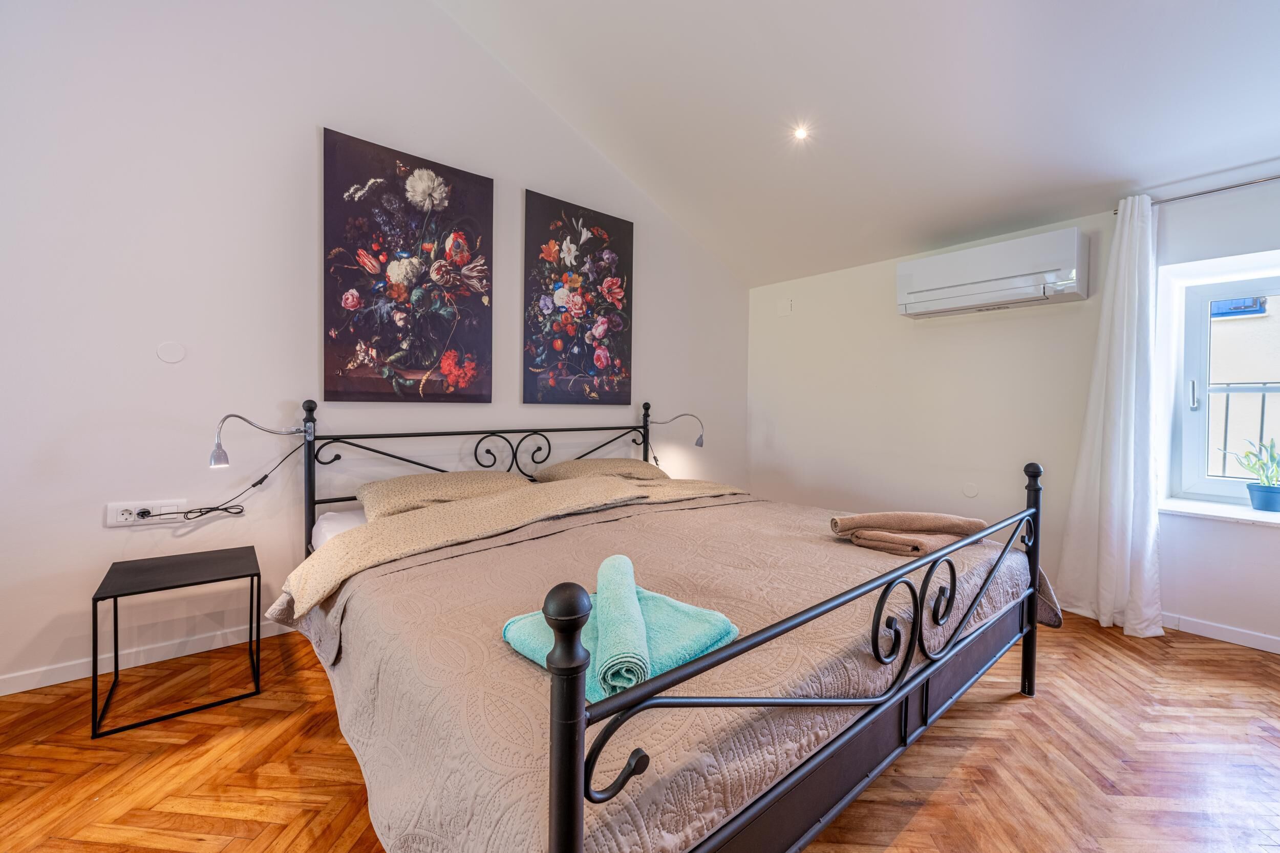 Ein Schlafzimmer mit einem Bett, einem kleinen Tisch, zwei Wandbildern, einer Klimaanlage und einem mit Vorhängen versehenen Fenster.