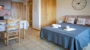 A bedroom with a bed, a wooden wardrobe, a small table with a chair, and a kitchen area in the background. - Seya 21 by Interhome (Ovronnaz)